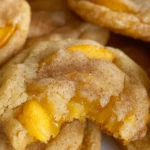 Three peach cobbler cookies on a ceramic plate with ripe peach and crumble in the background